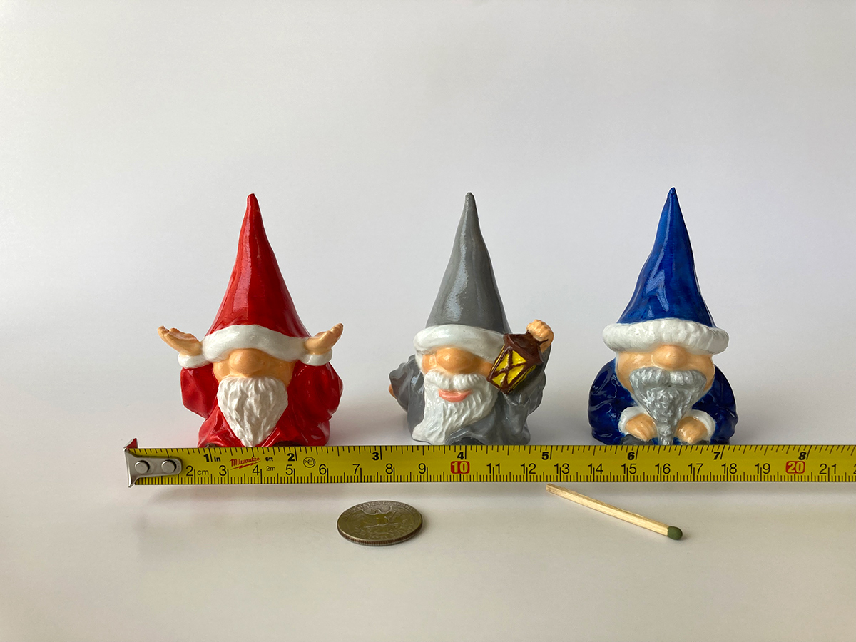 Hand-painted gnome figurines in red, gray, and blue hats placed next to a measuring tape for scale